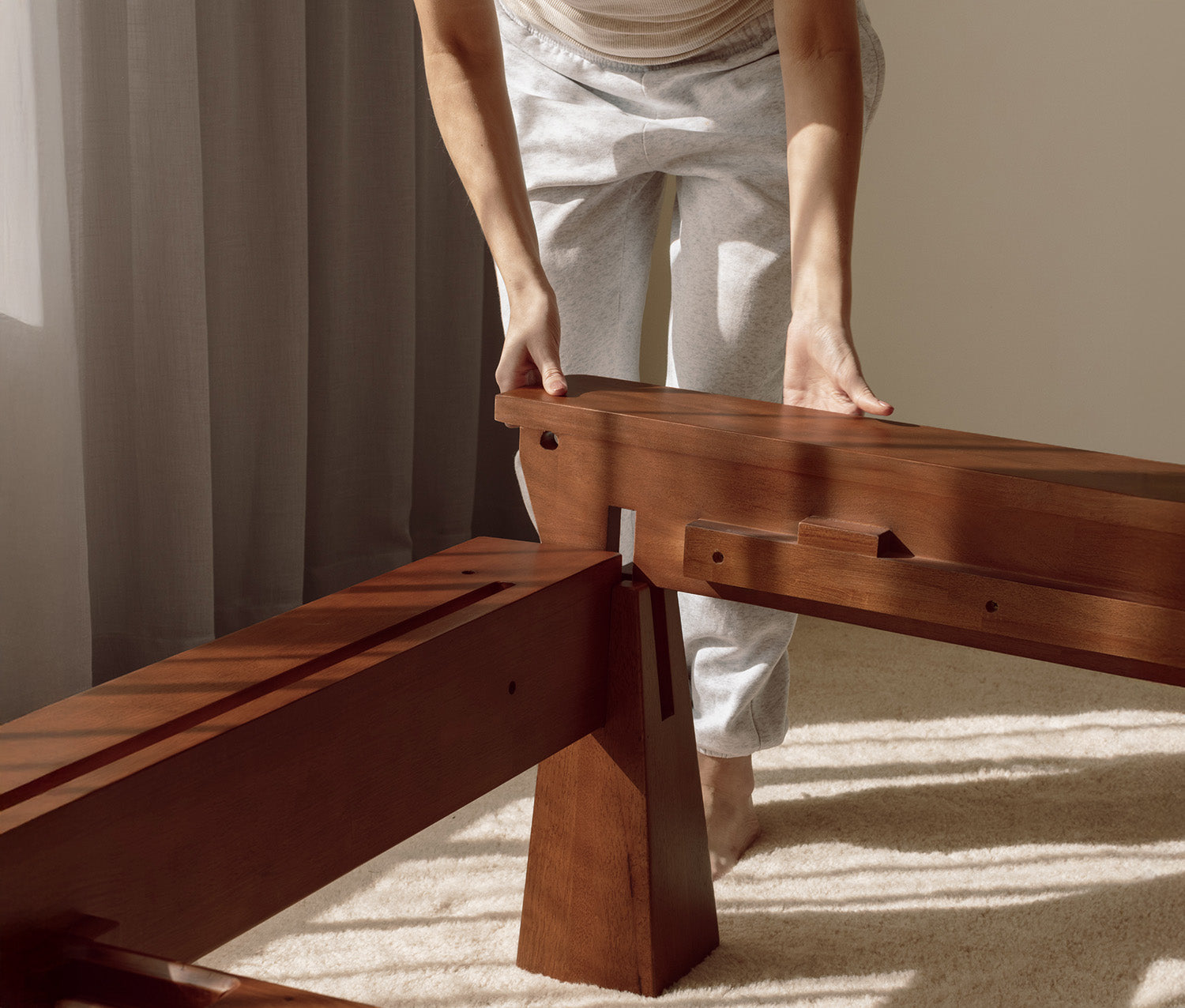 Person assembling a wooden bed frame with sunlight casting shadows on the floor.
