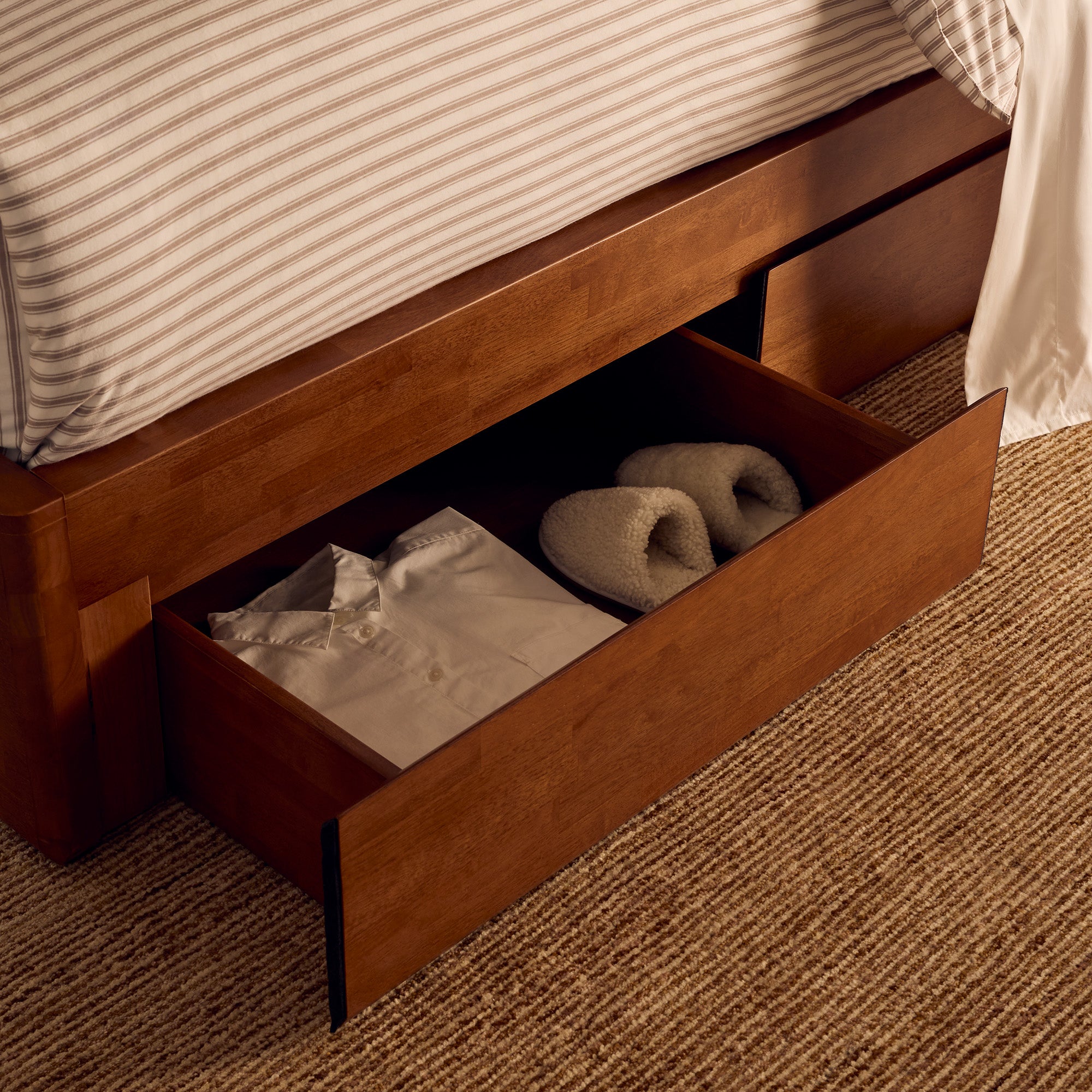 Wooden bed frame with open drawer containing folded clothes and slippers on a carpeted floor.